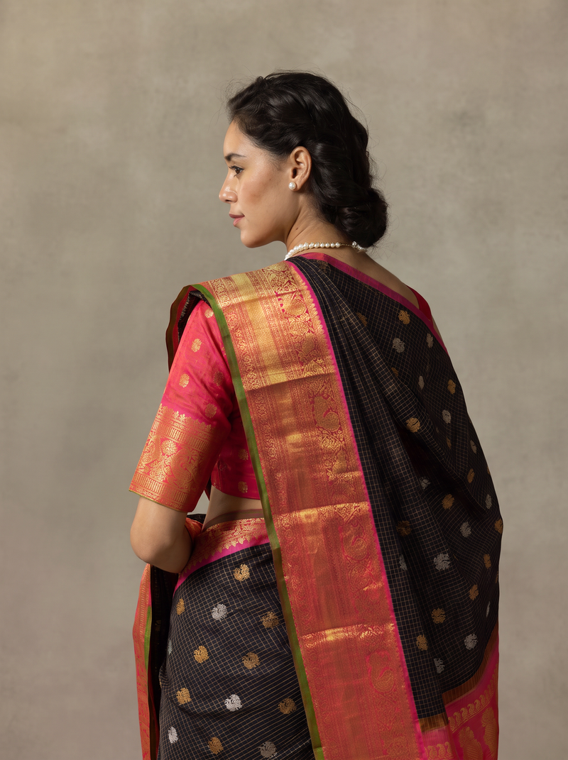 Gadwal - Handloom Gadwal silk in deep black zari checks with gold and silver butas. Its peach gold border and pallu glow with maroon pink paisleys and florals.