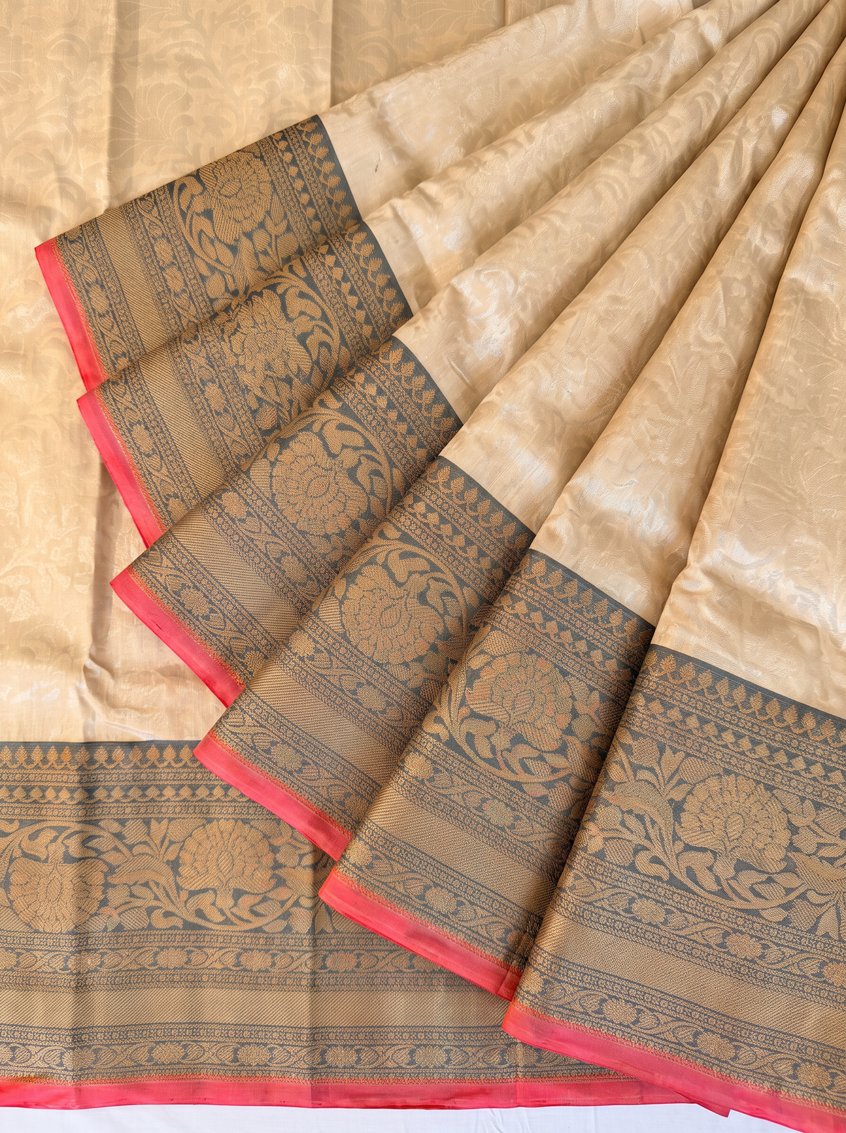 Like morning light on stone, soft cream meets warm grey, lit by a coral red edge and a shimmering gold zari jaal. Handloom Gadwal silk.