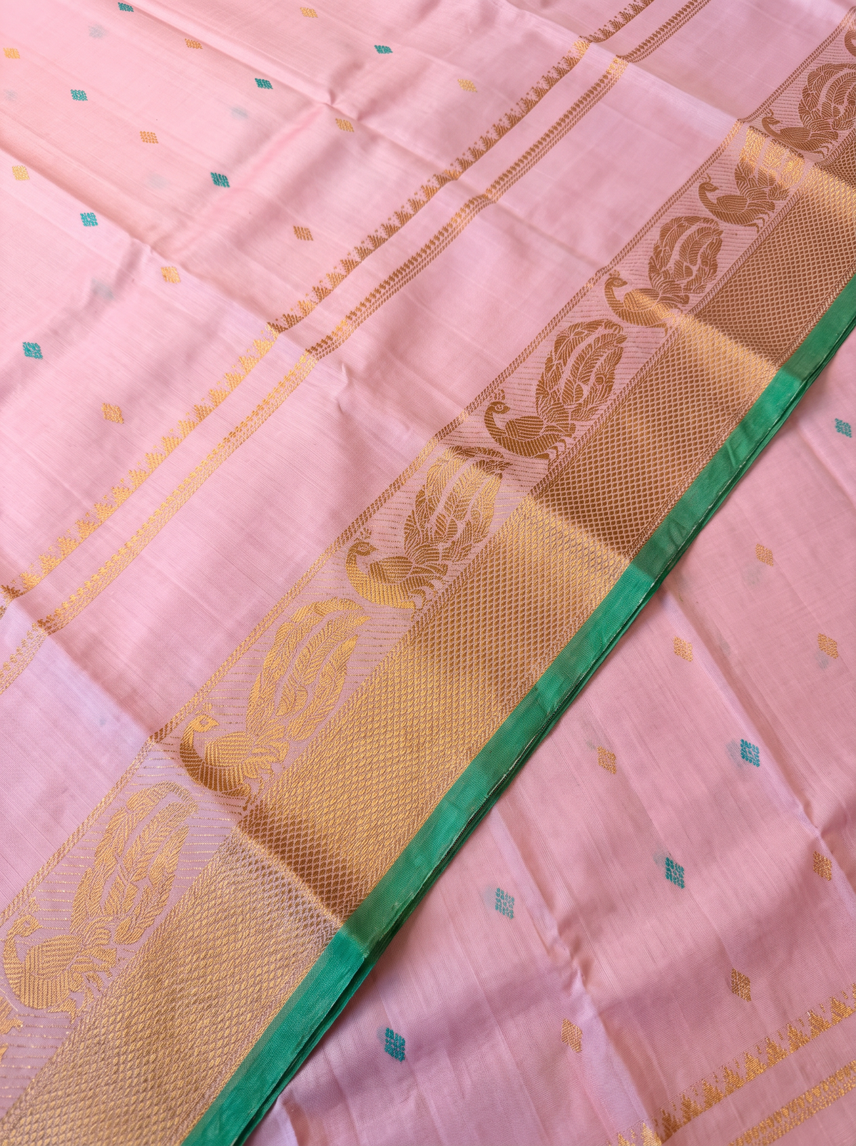 A blush of pink, a sprinkle of gold and teal, and peacocks in shimmering zari dancing along a green edge. Pure silk Gadwal, handwoven with love in Telangana.