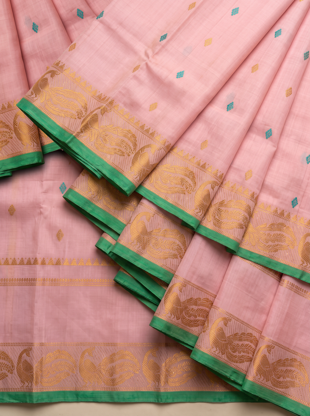 A blush of pink, a sprinkle of gold and teal, and peacocks in shimmering zari dancing along a green edge. Pure silk Gadwal, handwoven with love in Telangana.