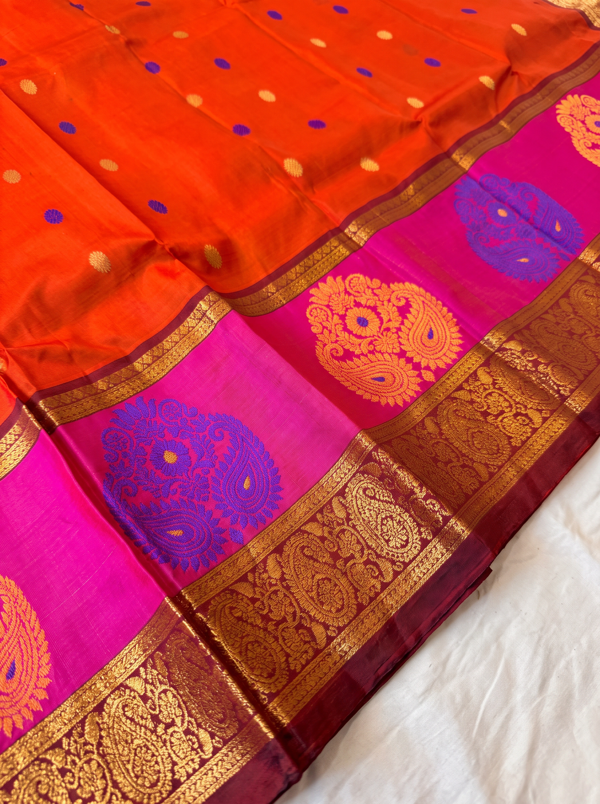 Like a marigold in full bloom, this orange Gadwal silk glows with gold and blue butis. A pink-edged maroon border holds it close, and the pallu shines in rich gold zari.