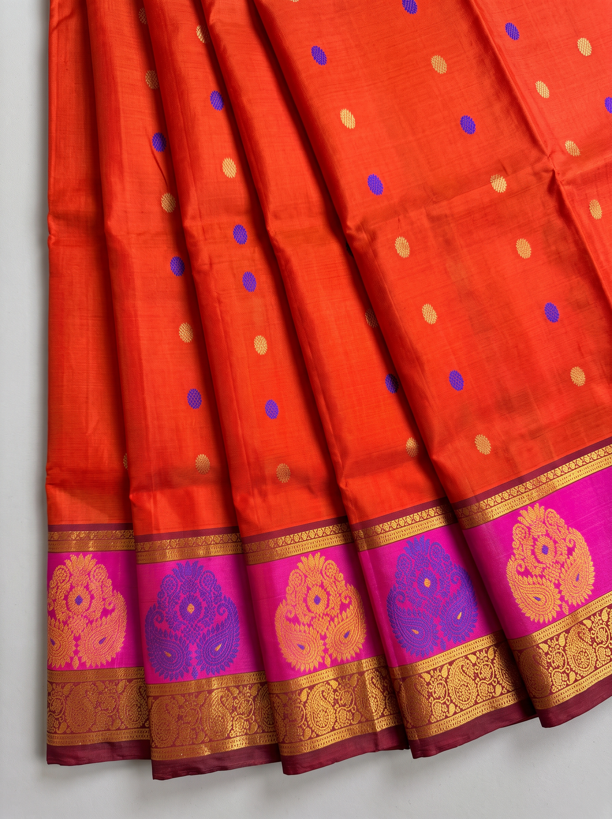 Like a marigold in full bloom, this orange Gadwal silk glows with gold and blue butis. A pink-edged maroon border holds it close, and the pallu shines in rich gold zari.