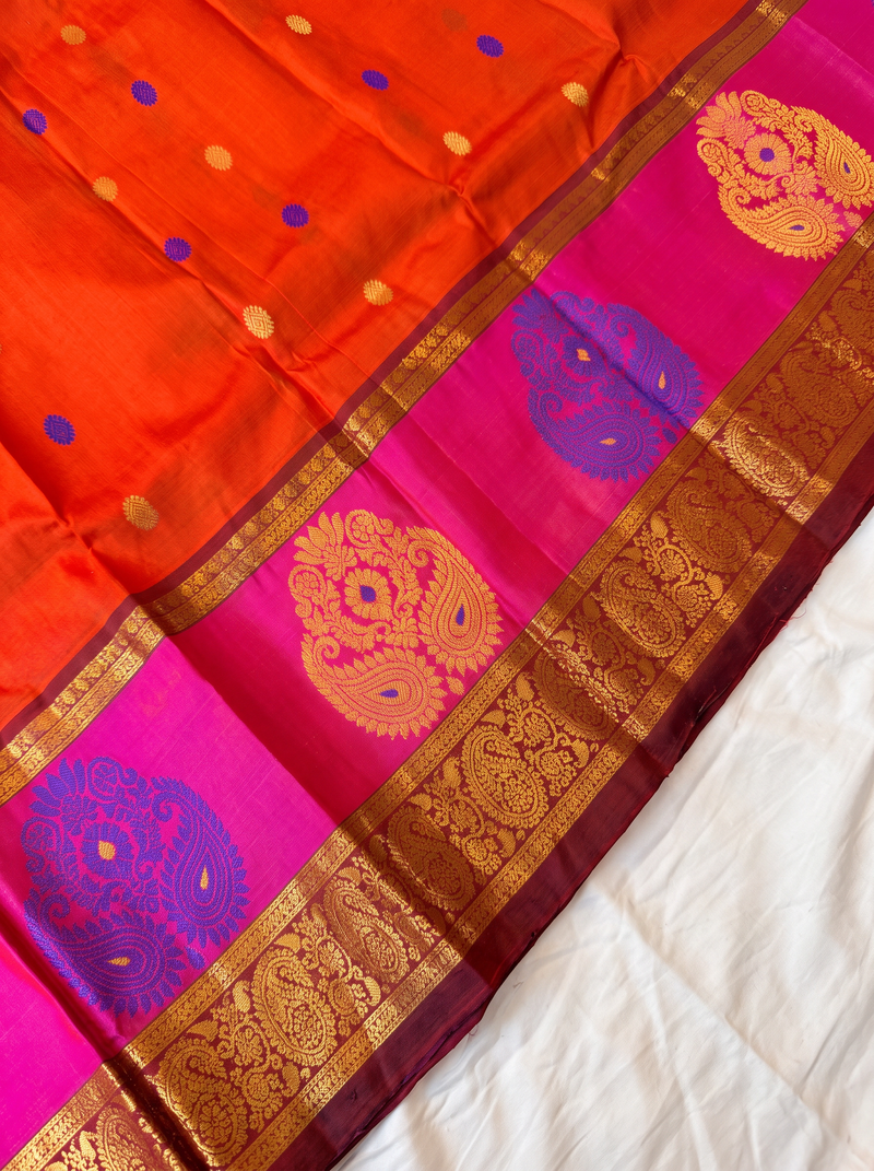 Like a marigold in full bloom, this orange Gadwal silk glows with gold and blue butis. A pink-edged maroon border holds it close, and the pallu shines in rich gold zari.