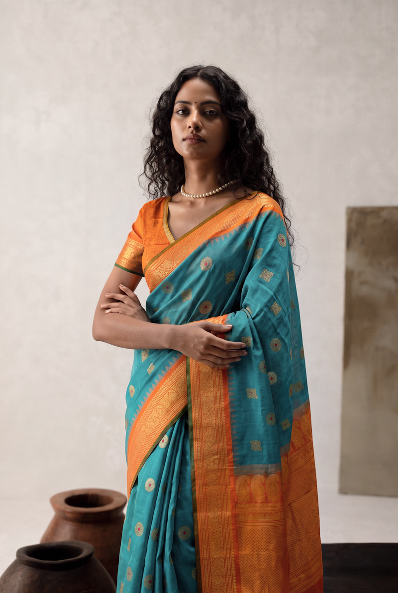 Rich teal, jewelled with ruby-dotted gold butas. A tissue-gold border that glows like liquid light, laced with diamond lattice and temple guards. Silk Mark certified handloom Gadwal silk from Telangana.